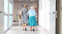 Older woman walking with cane down hospital hallway with younger woman