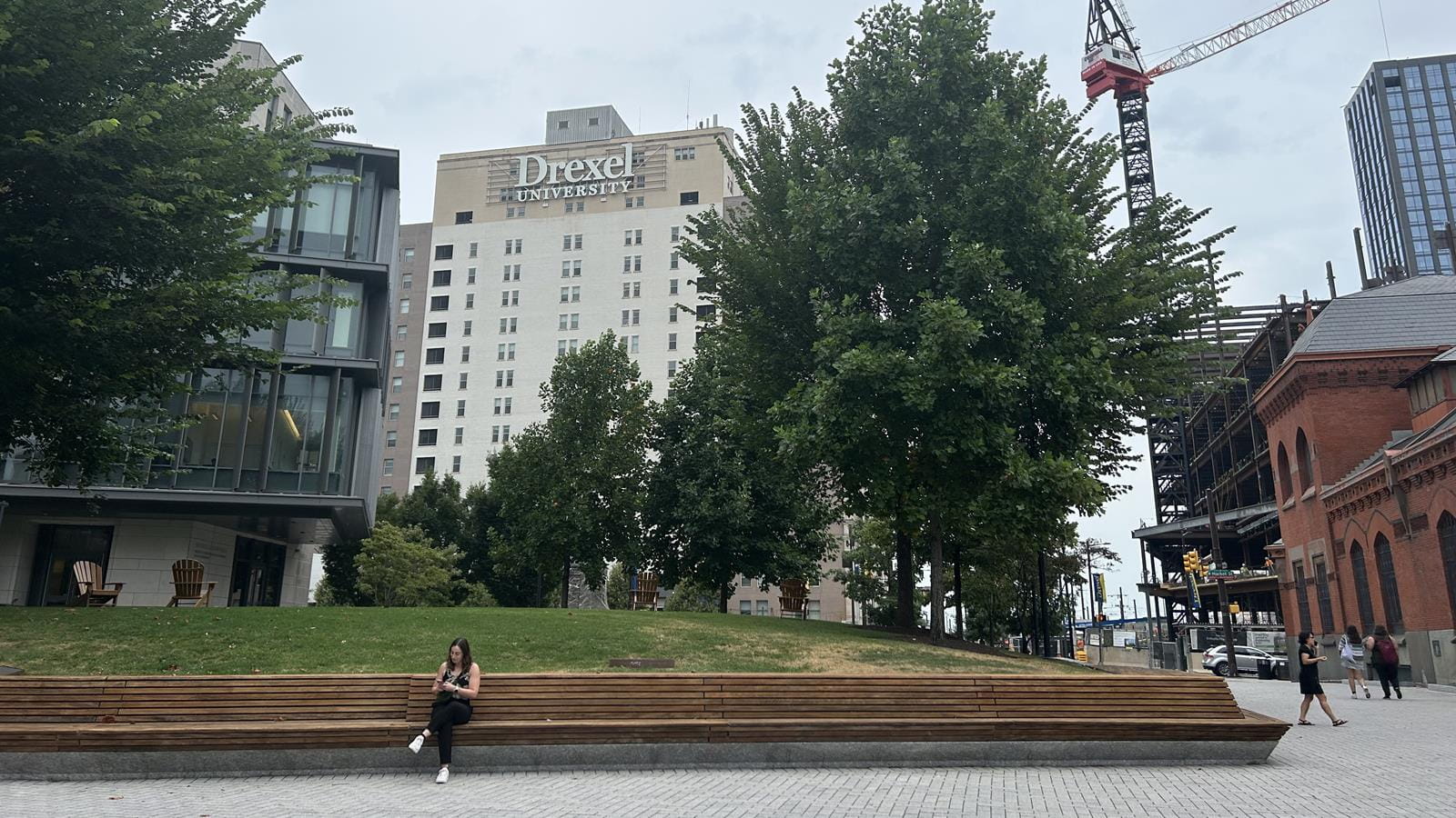 A woman sits on a brown bench in a paved outdoor area of Drexel University&#39;s Korman Quad with a construction crane in the background.