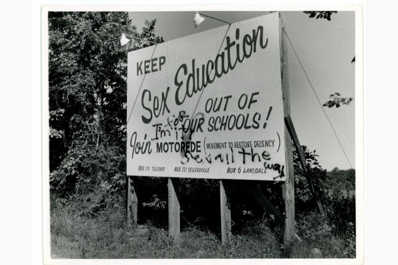 Road side sign advocating to "Keep Sex Education Out Of Our Schools" by the Movement to Restore Decency with locations in Telford, Sellersville, and Landsdale. A message spray painted over the sign reads, "I'm for it sex all the way." "One school of thought in the past," written on the back of the photo.