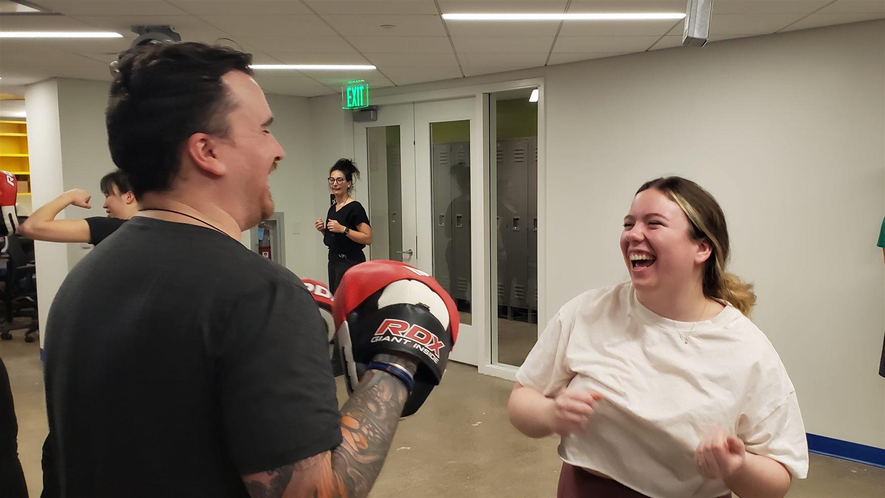 man in a black tee shirt with boxing gloves stands before a smiling female student in a white shirt who is laughing. as students look on.