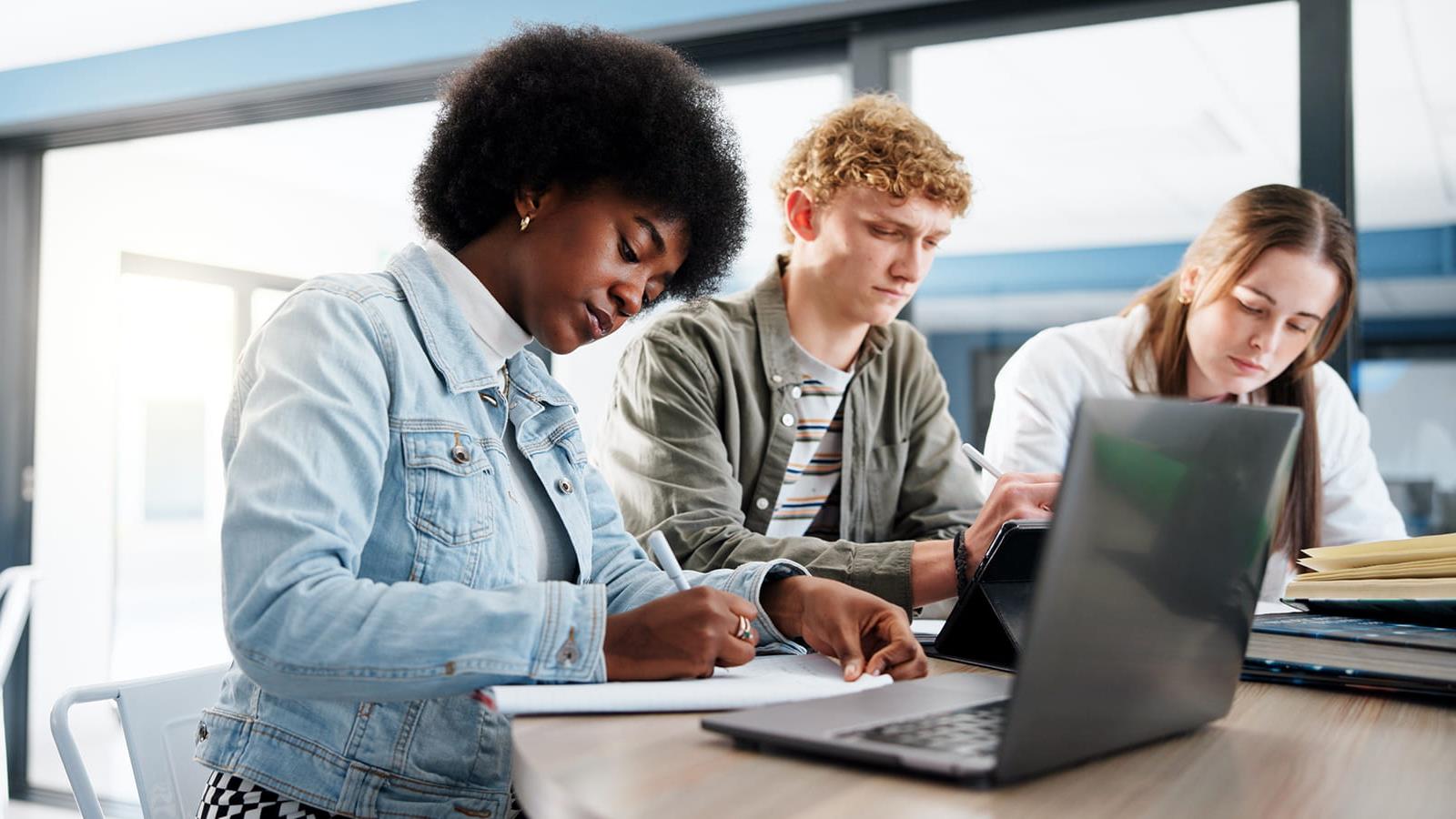 Three students sit in front of a lap top  writing down notes